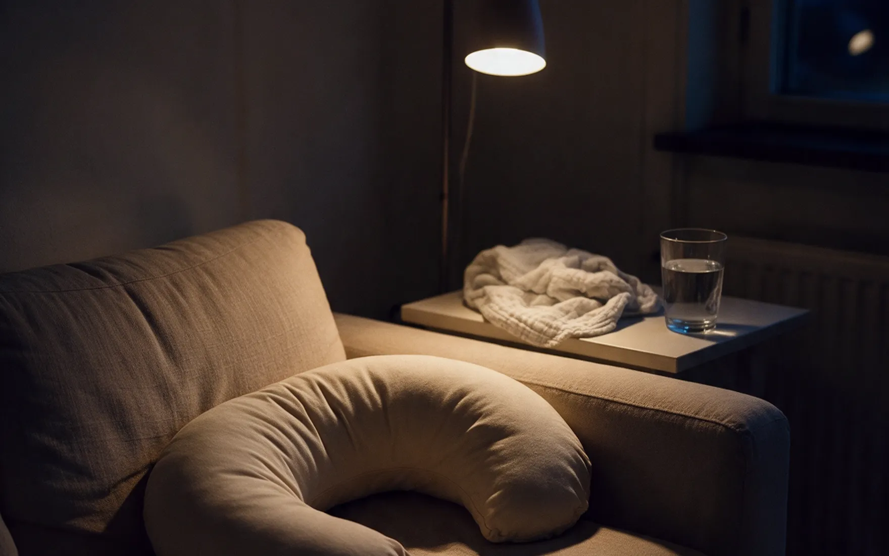 En amningskudde i soffan i kvällsljus, med ett glas vatten och en filt på bordet bredvid.